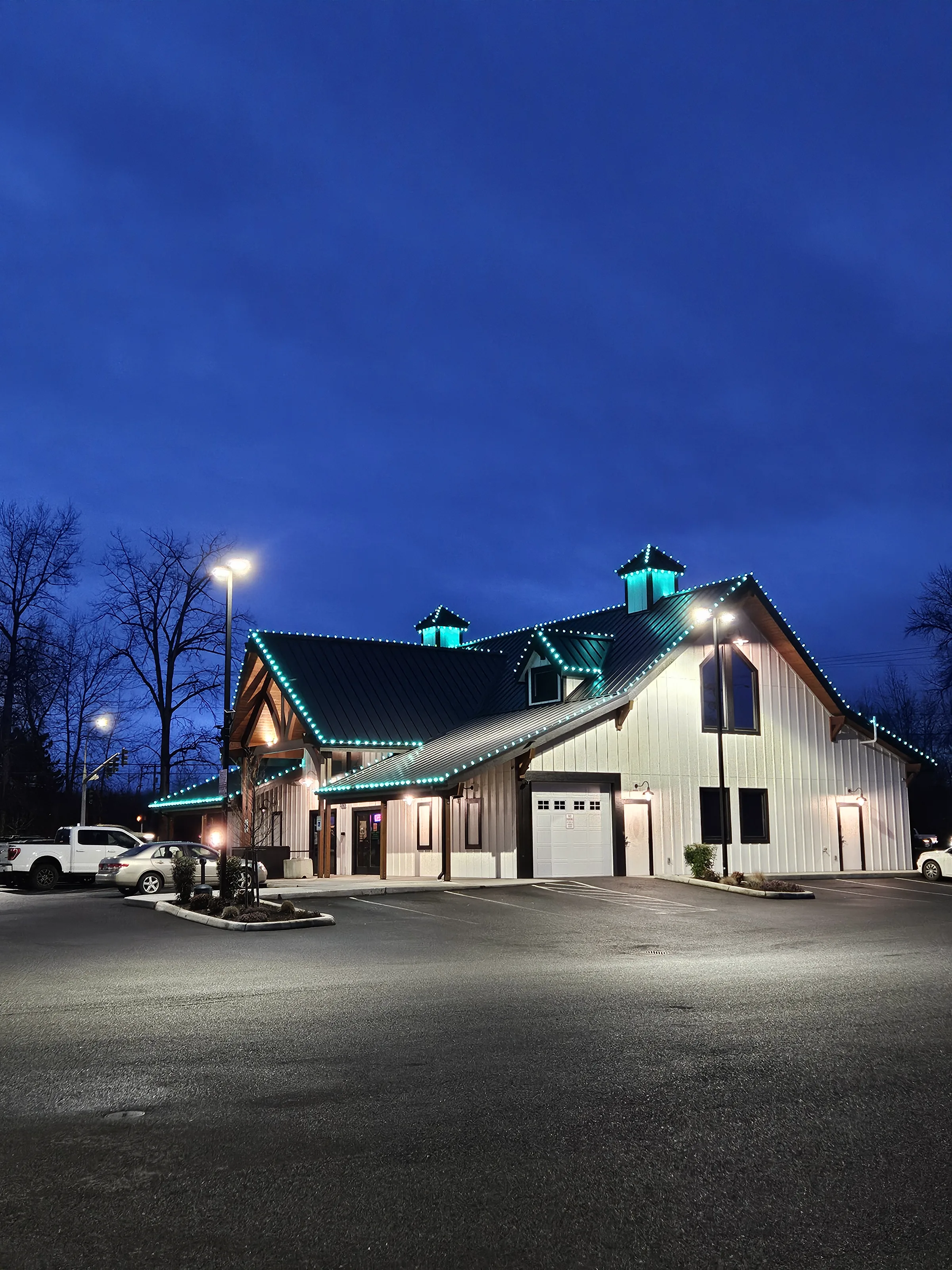 The Green Door at dusk: a white board-and-batten barn with a black standing-seam roof and two cupolas, its rooflines lit green, against a deep blue sky.